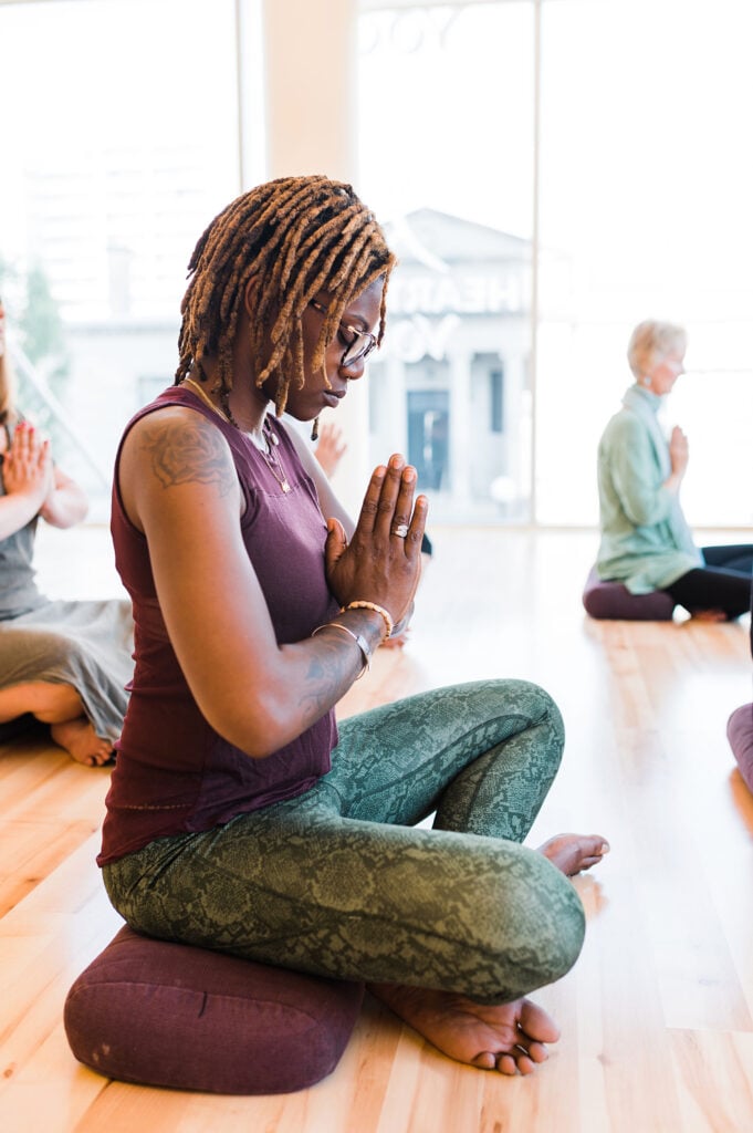A woman with brown skin sits cross-legged on a yoga mat with hands in prayer position in the Heartland Yoga Studio. In the background are two other yoga students seated cross legged.